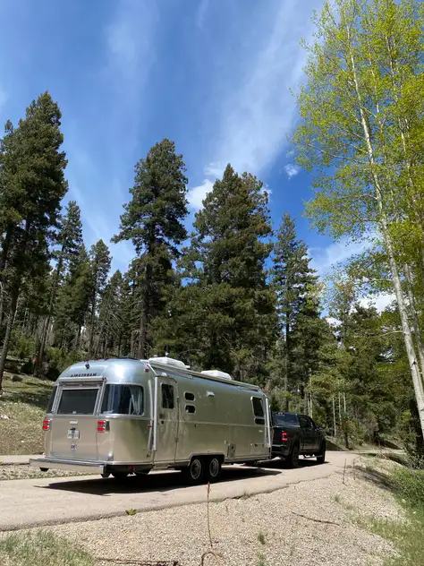 Airstream travel trailer parked on a paved pad surrounded by ponderosa pines and aspens under blue sky