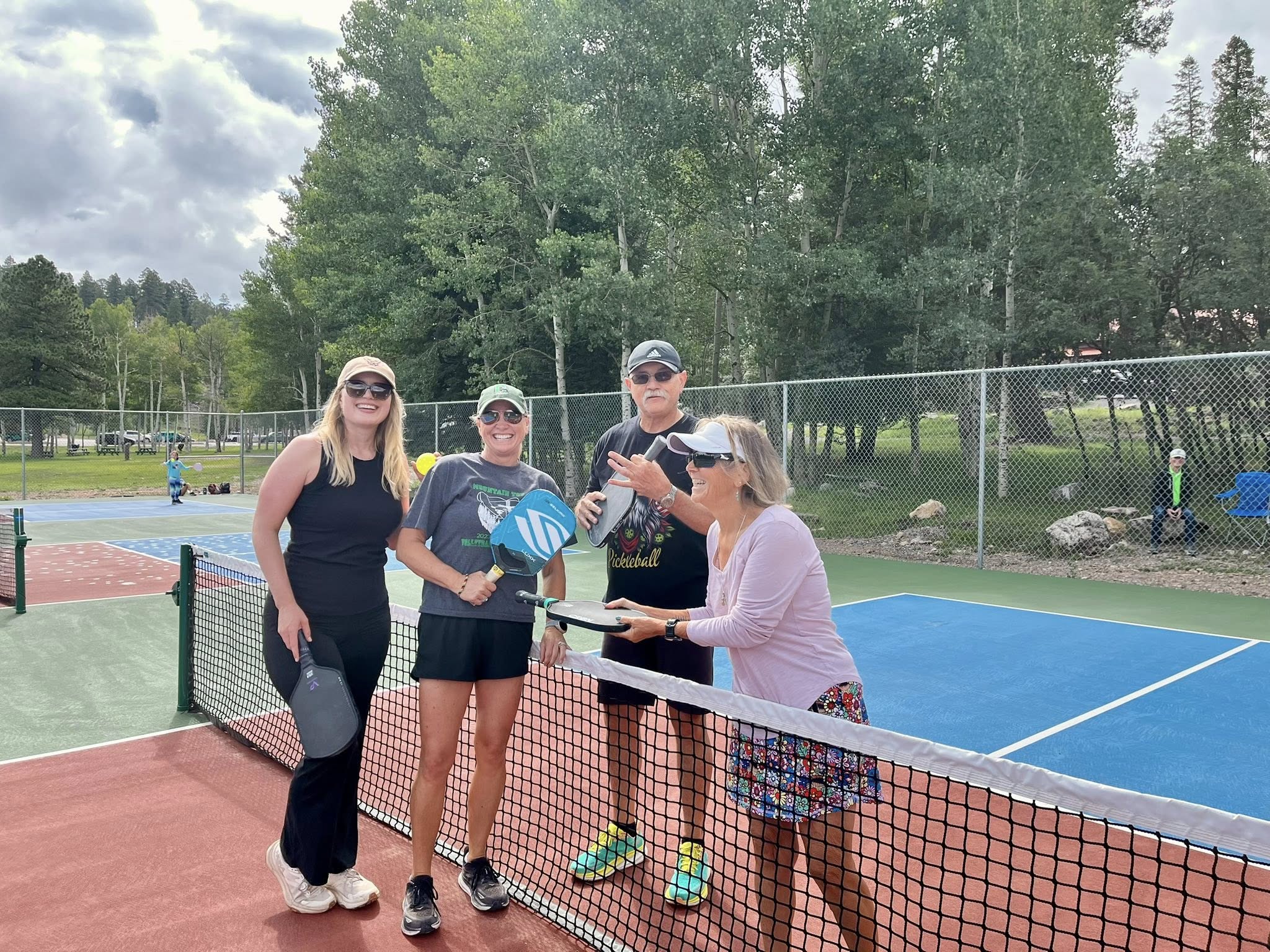 Four pickleball players pose at the net on Zenith Park courts with mountain forest behind them