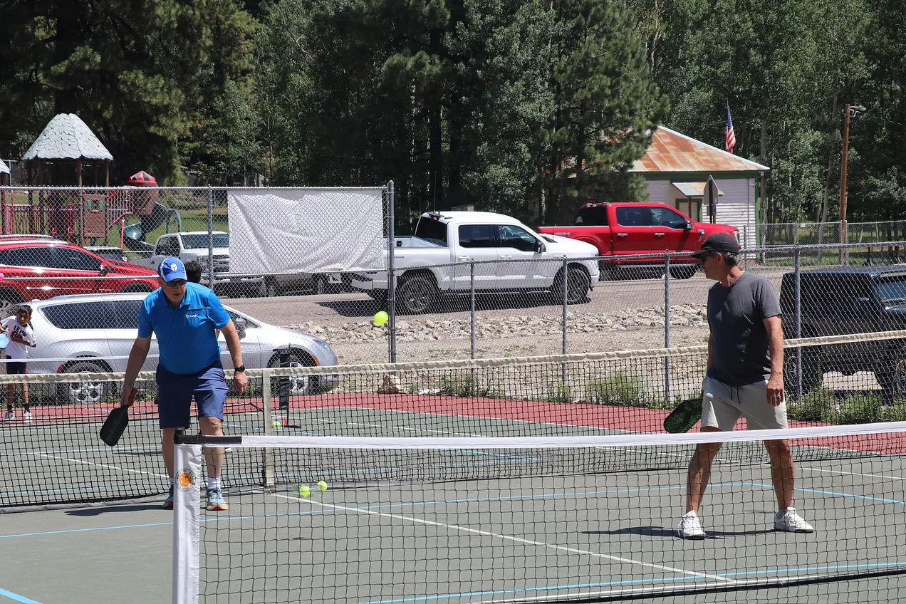 Two players at the net during a match with Zenith Park pavilion and pine trees in background