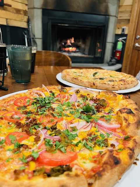 Two wood-fired pizzas on a wooden table in front of a stone fireplace with a live fire