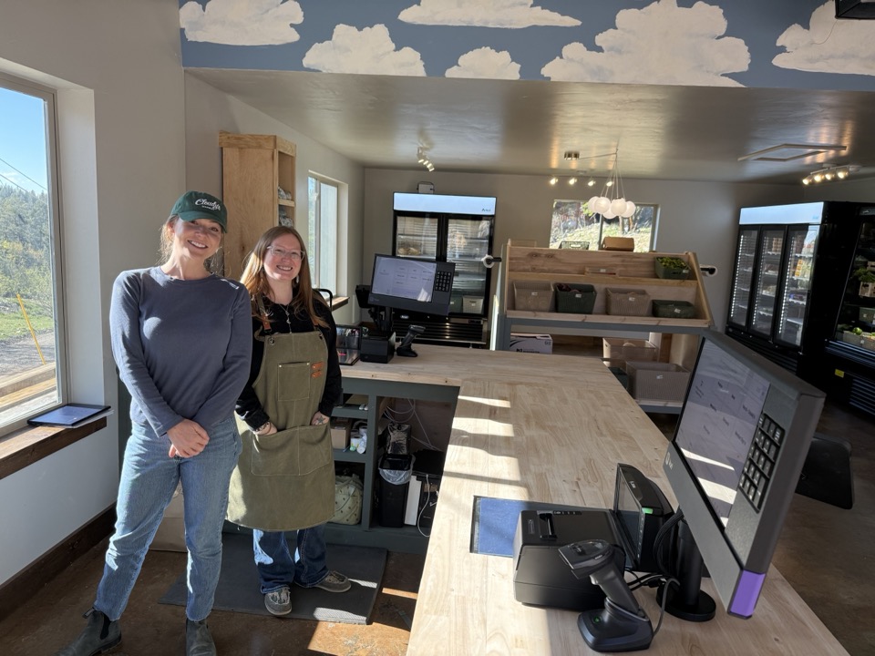 Two staff members behind the wood front counter at Cloudy's Market, with the painted clouds-on-blue ceiling and refrigerated coolers visible behind them