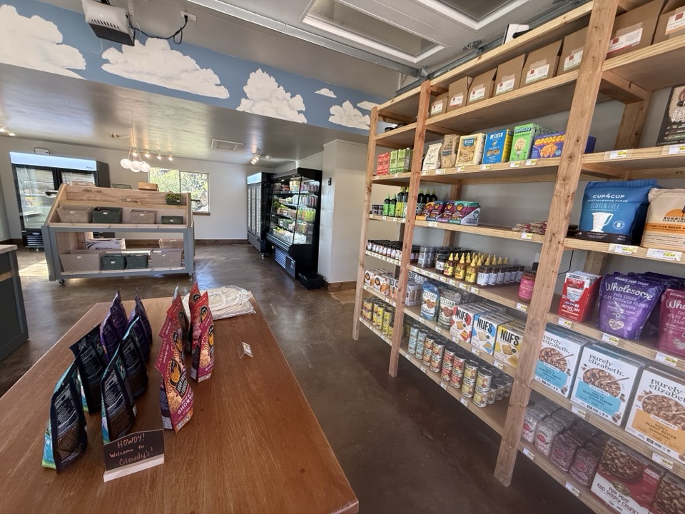 Wide interior view of Cloudy's Market — concrete floor, pine-shelved sales floor on the right, refrigerator case at the back, and clouds-on-blue mural across the front-of-house ceiling