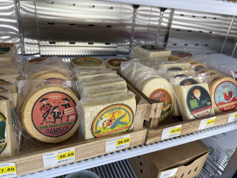 Wedges and rounds of Cheese Brothers cheese — including 3 Amigos jalapeño-habanero-chipotle gouda and green-onion cheddar — displayed in a cooler at Cloudy's Market