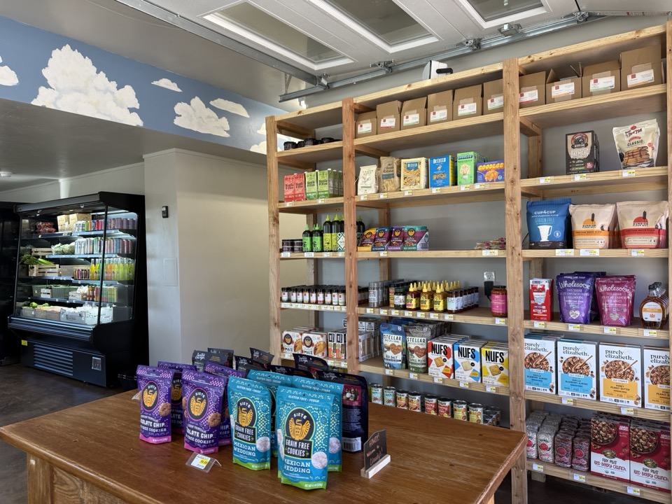 A wood table near the front of Cloudy's Market displays grain-free cookies and packaged snacks, with the pantry shelving and refrigerated case behind