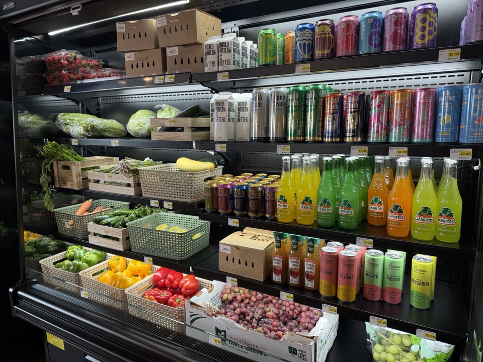 Refrigerated produce and beverage case at Cloudy's Market — bell peppers, tomatoes, grapes, leafy greens, and rows of bottled and canned drinks