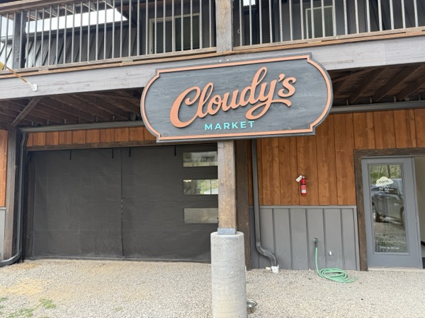 The Cloudy's Market sign above the entrance — a script logo on a charcoal panel mounted to the cedar-sided storefront on Mexican Avenue