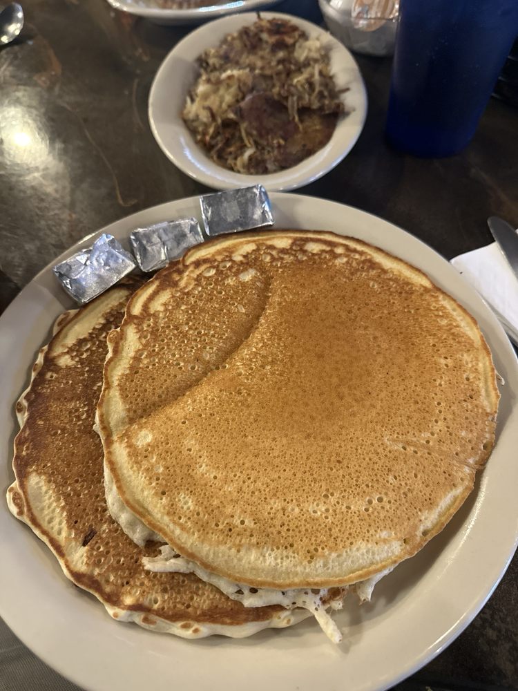 A stack of large pancakes with butter pats on the side and a bowl of hash browns