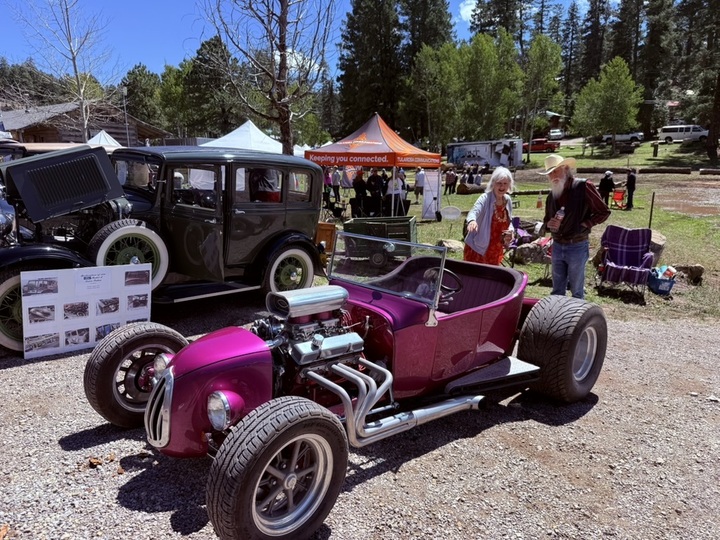 A magenta hot rod and a vintage black sedan parked among the pines at a Cloudcroft car show, with vendor tents and spectators behind them