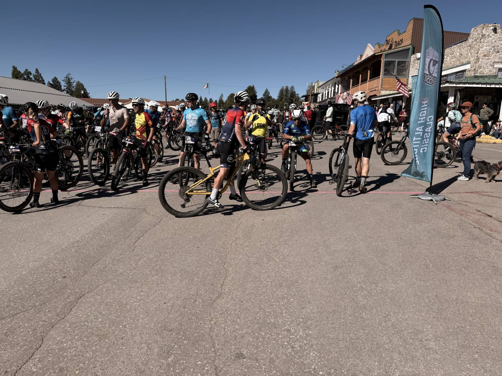 Wide start line at the High Altitude Classic — riders lined up in downtown Cloudcroft with the branded flag banner