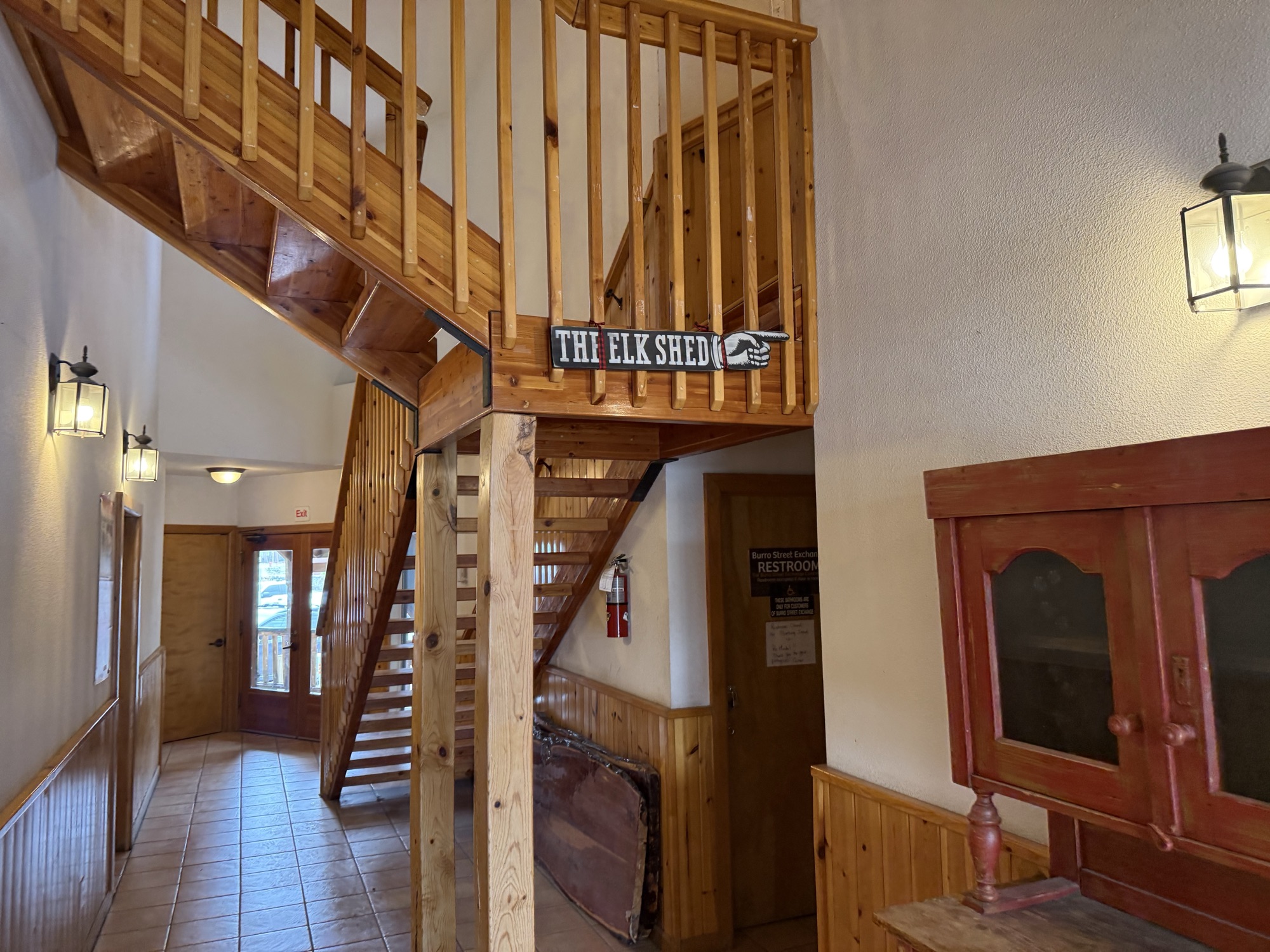 Open stairwell to the upstairs loft at The Elk Shed with pine-paneled walls, vintage stagecoach detail, a carved THE ELK SHED wood-plank sign with elk silhouette mounted on the loft railing, and a red glass-front display cabinet