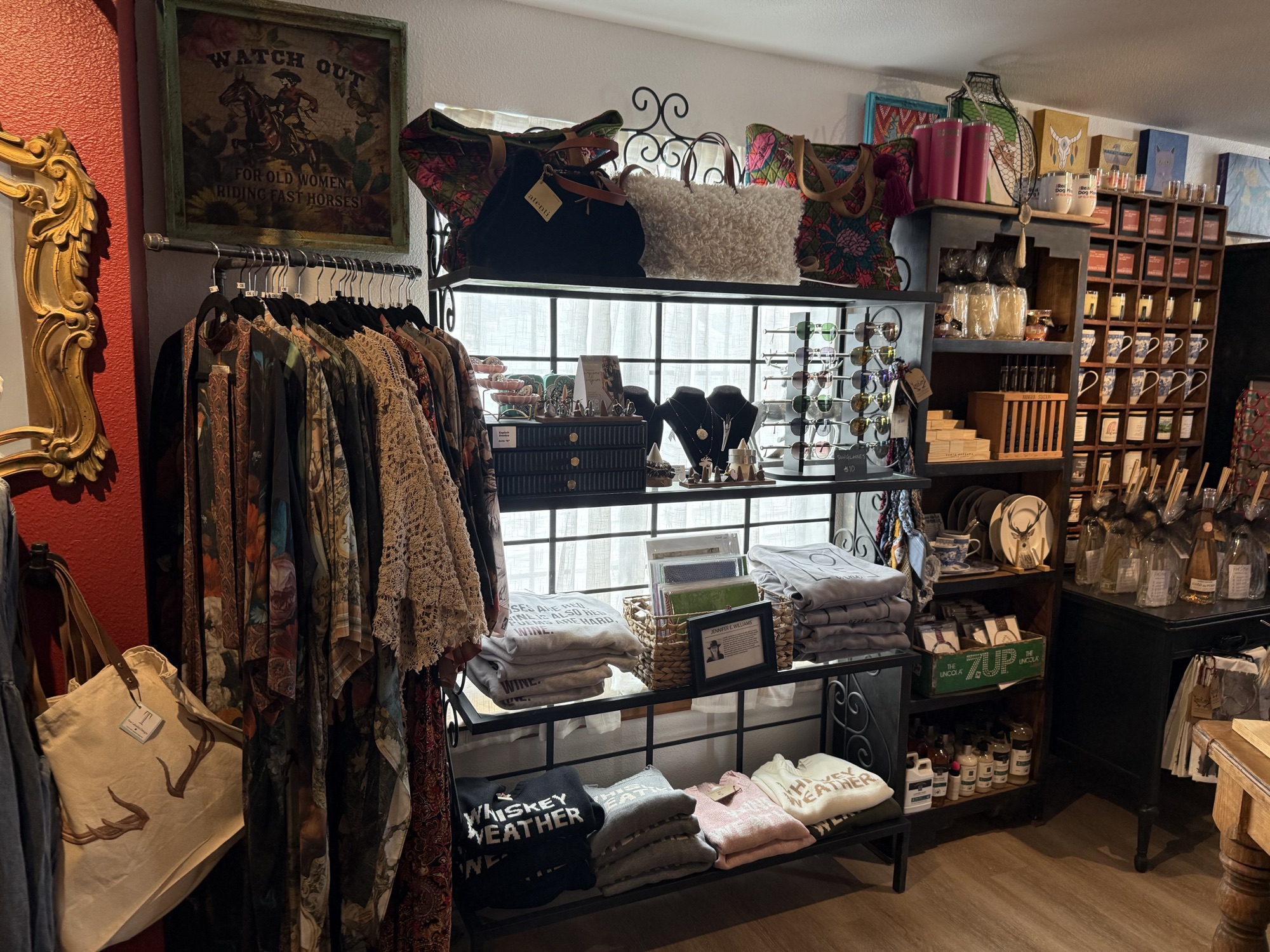 Gourmet-and-gift shelf at The Elk Shed with patterned tops on a clothing rack, Whiskey Weather tea towels stacked on shelves, candles, jars, and small jewelry on a black wire display