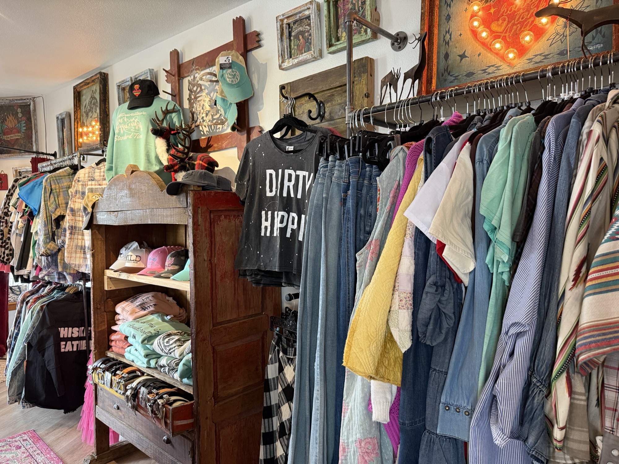 Clothing rack at The Elk Shed with a featured Dirty Hippie graphic tee, denim and printed skirts, framed Western art on the wall, and a hat shelf with small dolls and stacked tees