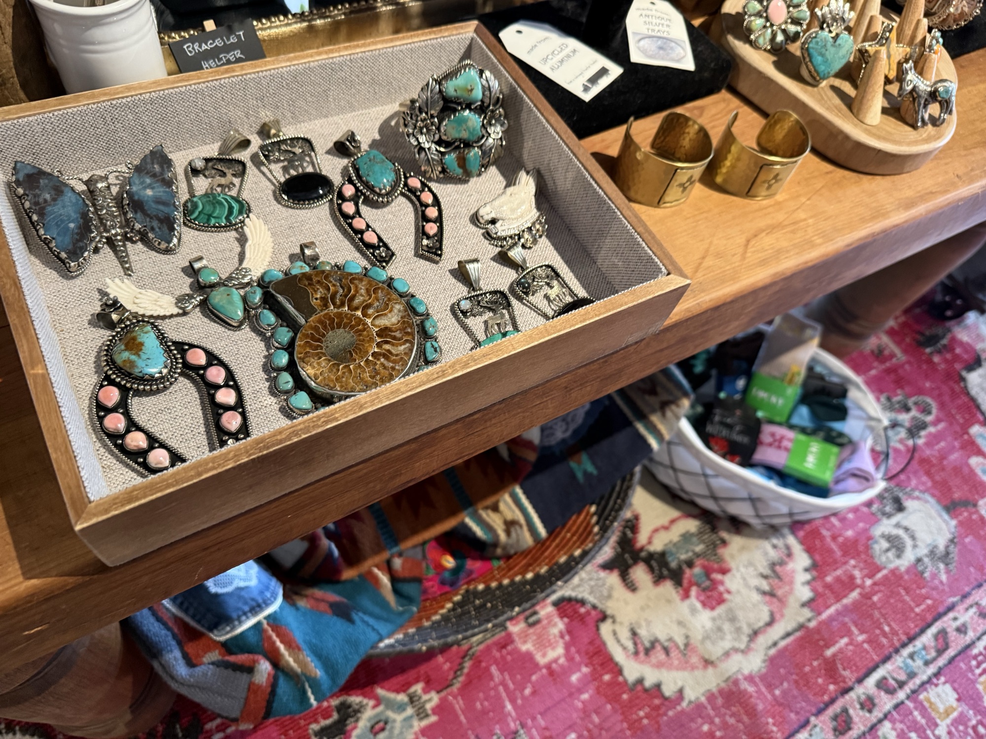 Closeup of a jewelry tray at The Elk Shed with turquoise-and-coral pendants, an ammonite pendant, butterfly pendant, gold and silver cuffs, and ring stands on a wooden table over a Persian rug