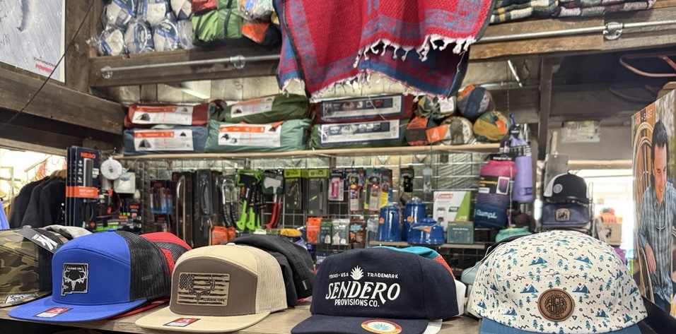 Hat shelf at High Altitude with Sendero, Snake Farm, and Hill Country caps above a row of fishing tackle and a Southwest blanket