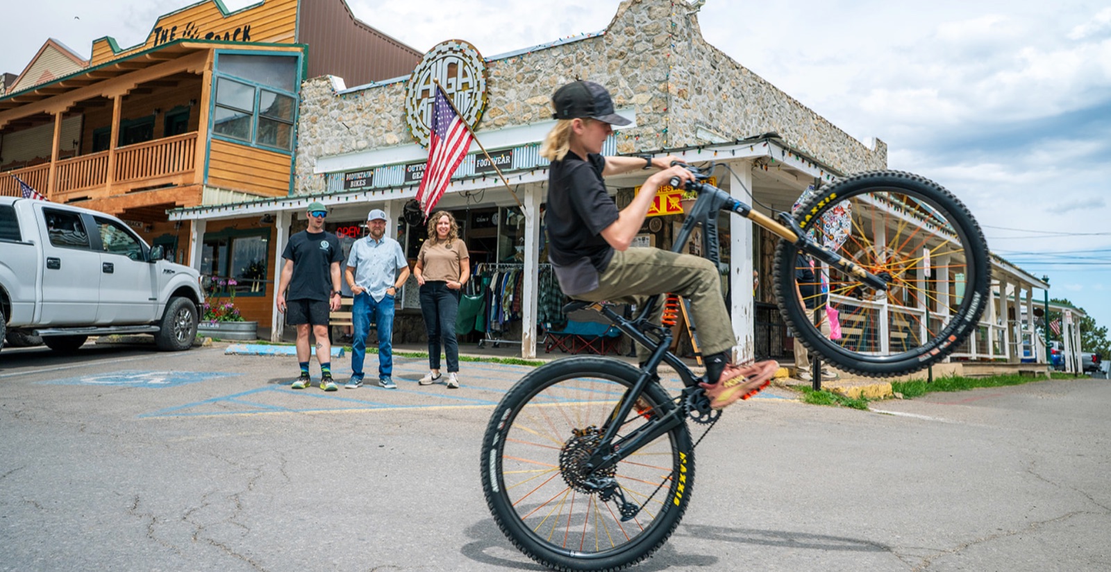 Miles Willett rides a mountain bike wheelie past the stone High Altitude storefront on Burro Avenue while three staff members watch from the porch