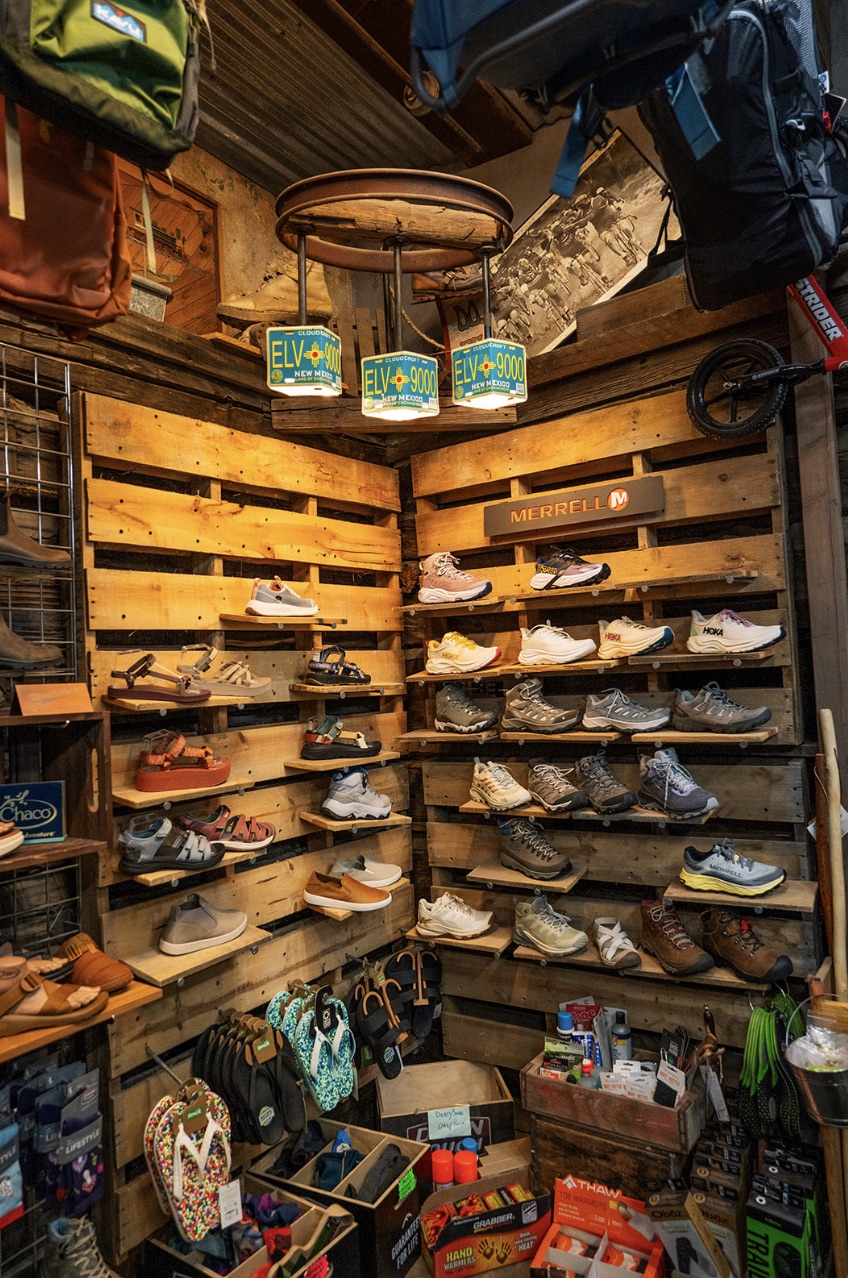 Corner footwear wall at High Altitude with Merrell, Hoka, and Chaco shoes and sandals on pallet shelving under Cloudcroft ELV 9000 license-plate lamps