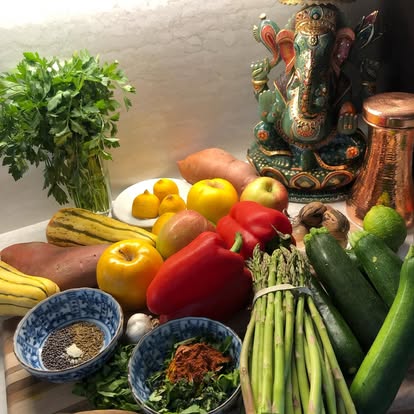 Fresh produce arranged with a Ganesh statue at Instant Karma — peppers, asparagus, sweet potato, citrus, herbs in spice bowls