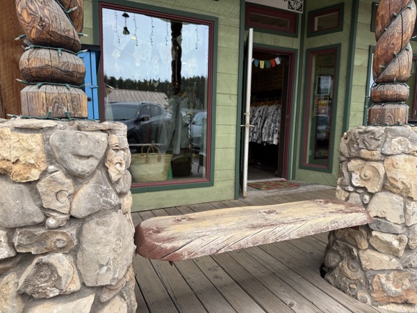 Stone-column porch at Instant Karma with carved wood figures, wooden bench, and view through the front window into the shop