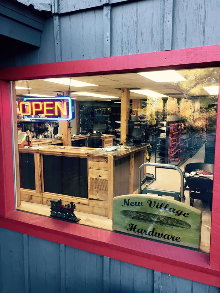Red-framed front display window at New Village Hardware with neon OPEN sign, lit interior, and a New Village Hardware wooden sign on the counter beside a train figurine