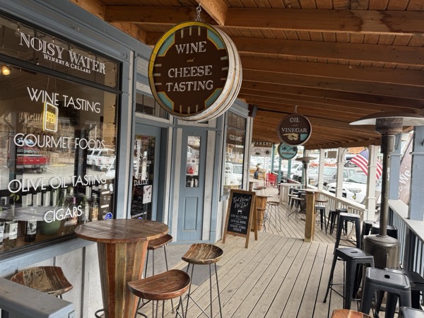Noisy Water Winery — boardwalk porch view at the Burro Street Exchange with the painted 'Wine and Cheese Tasting' barrel-end sign and the WINE TASTING / GOURMET FOODS / OLIVE OIL TASTING window menu