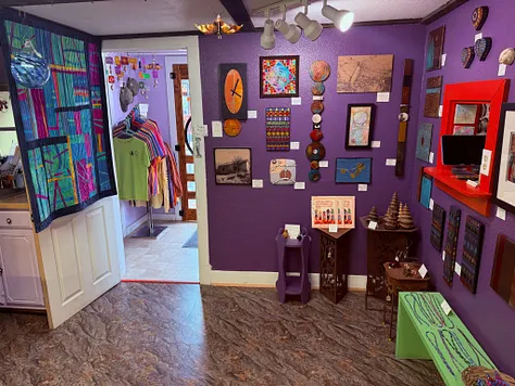 Purple-walled gallery room with hanging quilts, framed art, a clock, and wooden display tables