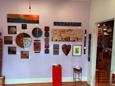 Mixed-media wall display &mdash; landscape paintings, heart-shaped collages, and painted wood discs on a lavender wall