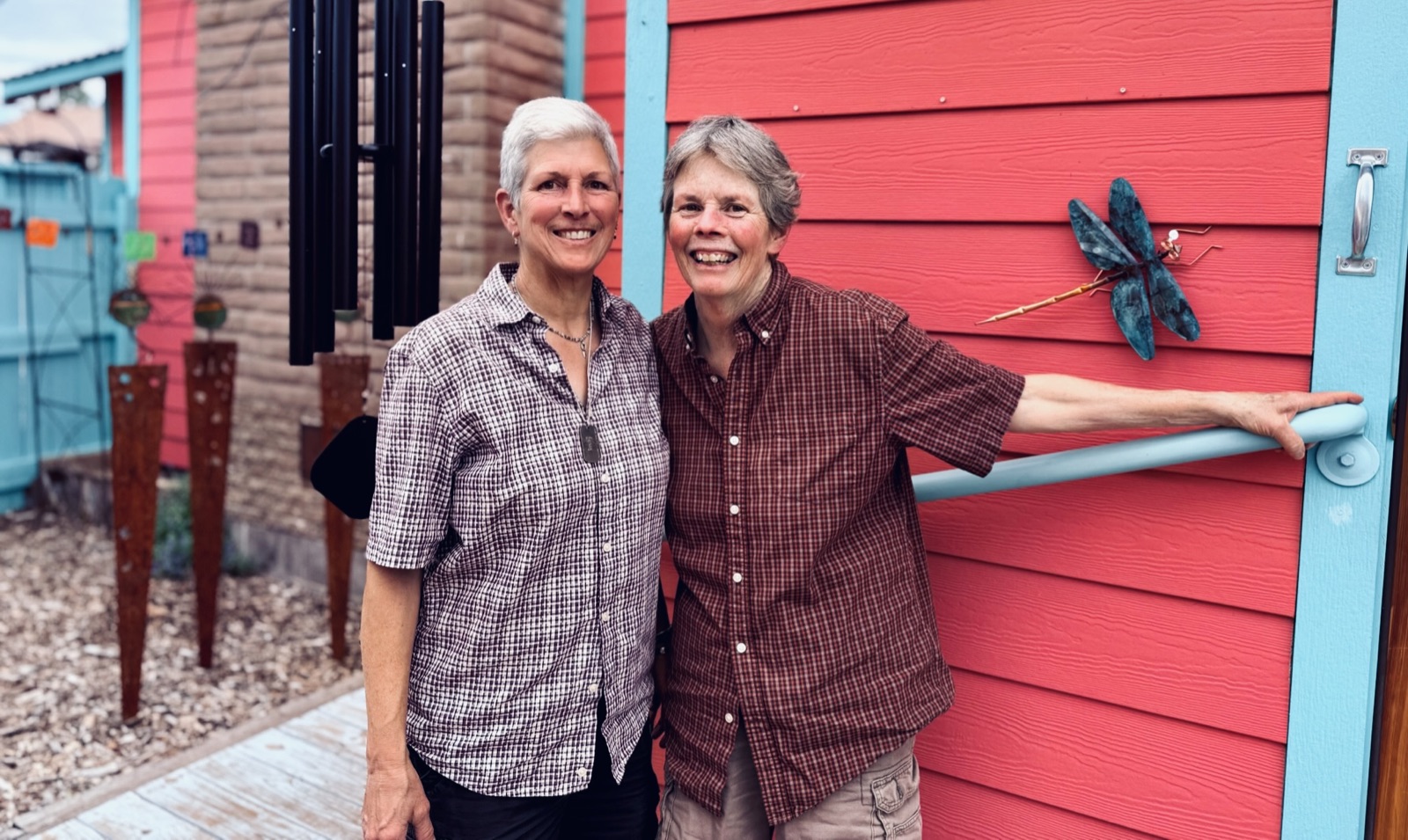 Berle Van Zandt and Donna Rand stand together in front of the pink-and-turquoise gallery door, beside a verdigris metal dragonfly and a set of black wind chimes