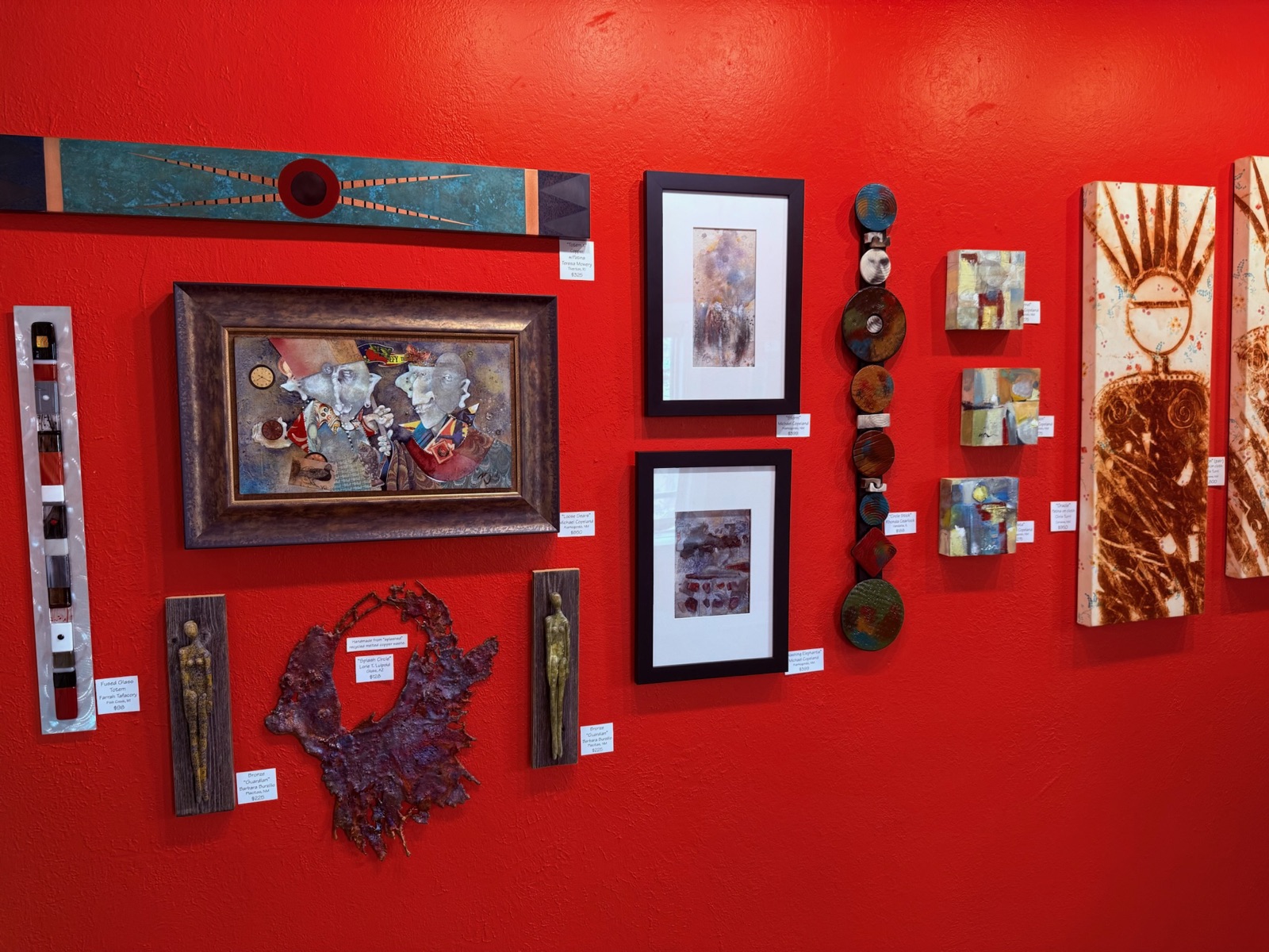 Red gallery wall hung with labeled original art — framed Michael Copeland paintings, a copper totem, bronze figures, and Lorie T. Luipold's recycled-metal Splash Circle