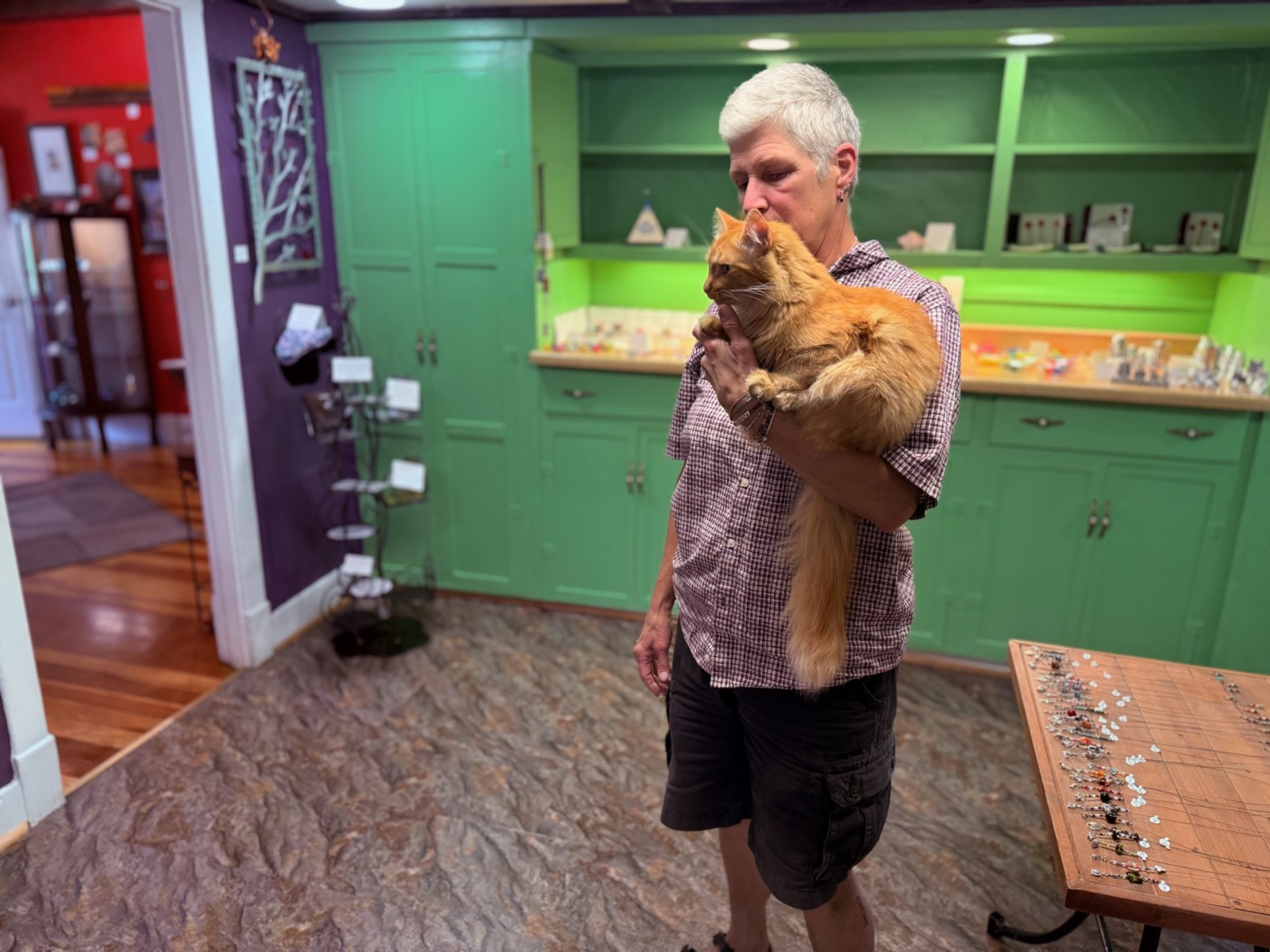 Berle Van Zandt holds Jules, a long-haired orange cat, against her shoulder in a green-cabinetted gallery room lined with jewelry displays