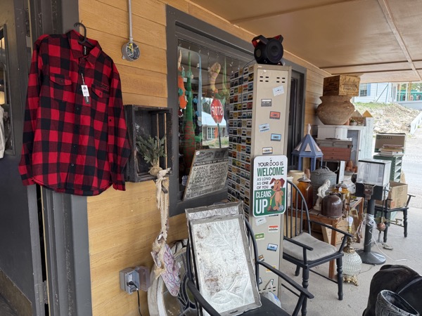 Front porch detail at Poke The Bear with red plaid flannel hanging by the door, a Your dog is welcome sign, antique chair, and stacked frames