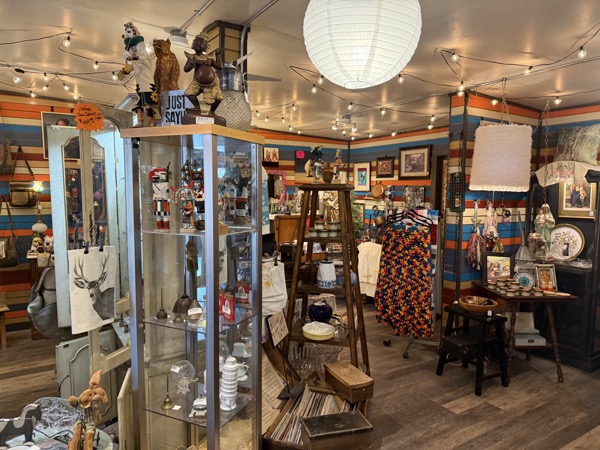 Vintage and antique back room at Poke The Bear with carved figures, ladder display, mannequin in a printed dress, and orange-and-blue striped walls strung with cafe lights