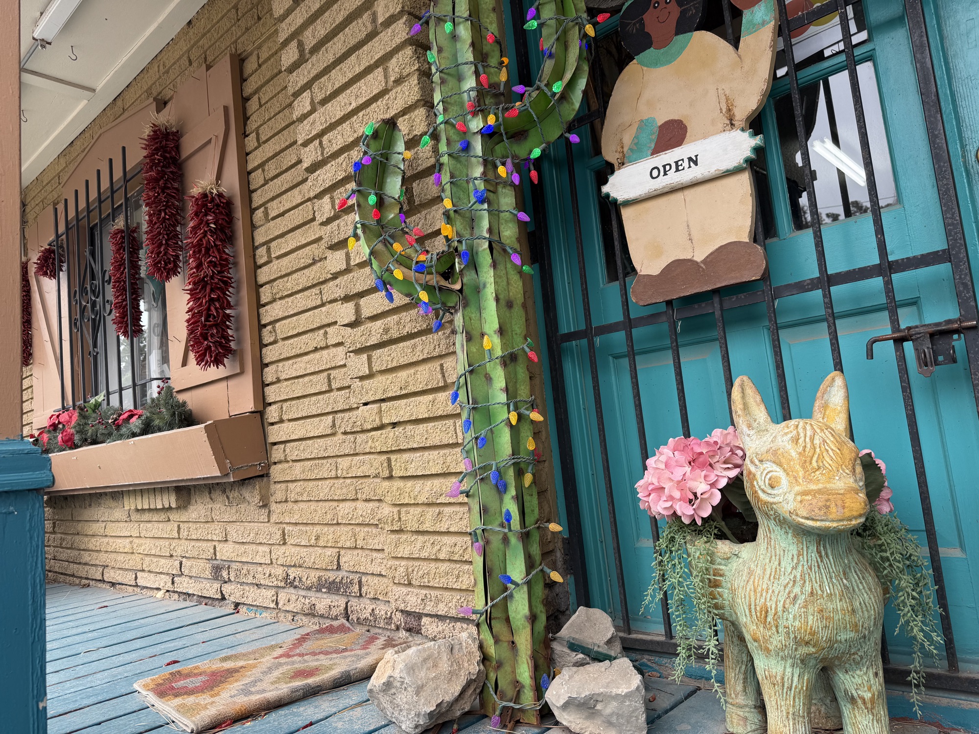 Front entrance of The Turquoise Shop with ristras, holiday-lit cactus, OPEN sign, and a hand-painted donkey planter