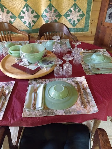 Breakfast table at Burro Street Boardinghouse set with green jadeite glassware, hand-stitched quilt on the wall, and antique placemats